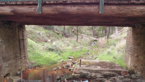 Drone-flys-toward-and-under-a-timber-and-stone-bridge-over-a-stream,-with-a-weaterboard-shed-on-the-left,-in-the-Australian-bush