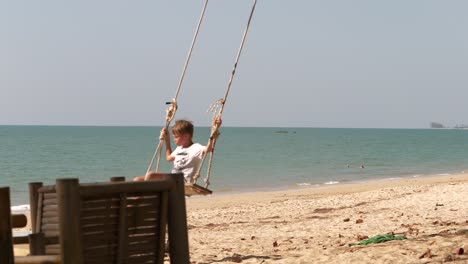 Boy-on-a-swing,-at-the-Pak-Weep-Beach,-the-Andaman-sea-in-the-background,-sunny-day,-in-Khao-Lak,-Thailand---slow-motion