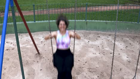Young-woman-with-long-skirt-and-colorful-shirt-swinging-and-laughing-feeling-free-and-happy-at-a-park-viewed-from-the-front-with-fixed-camera