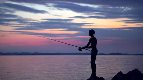 Silhouette-Of-A-Shirtless-Young-Man-Standing-On-The-Rocks-Near-Sea-While-Trying-To-Catch-A-Fish-During-Sunset---Wide-Shot