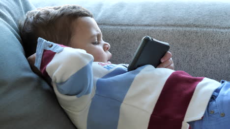 Kleiner-Junge,-Der-Auf-Der-Couch-Liegt-Und-Ein-Smartphone-Hält,-Das-Filme-Von-Der-Seite-Ansieht