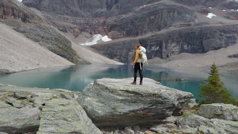 A-woman-hiker-stands-on-a-huge-boulder-taking-in-the-majestic-reflections-of-Oesa-Lake,-Alberta,-Canada