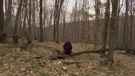 Woman-in-purple-jacket-with-backpack-resting-on-a-fallen-tree-in-Hoia-Baciu-forest-in-Romania-in-winter