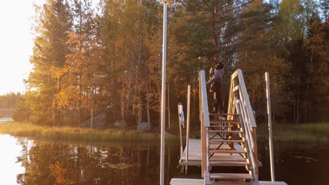 Boy-standing-on-wooden-bridge-enjoying-golden-lake-sunset-and-orange-autumn-colors,-in-Ostrobothnia,-Finland