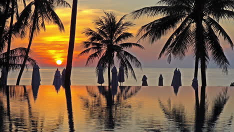 Static-shot-of-an-infinity-pool-during-sunset,-showing-the-orange-colored-sky-and-a-silhouette-of-palm-trees,-beach-umbrellas-and-the-ocean-in-the-background