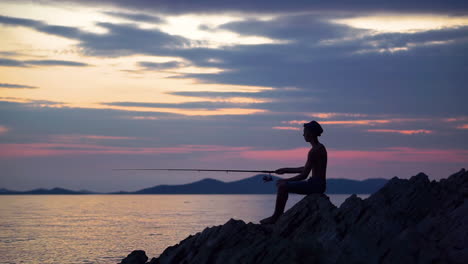 Young-Man-Wearing-Hat-Sitting-On-The-Rocks-Near-Sea,-Trying-To-Catch-Fish-During-Sunset---Wide-Shot