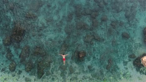 Young-Caucasian-man-swims-out-from-the-shore-into-open-turquoise-water,-high-overhead-aerial