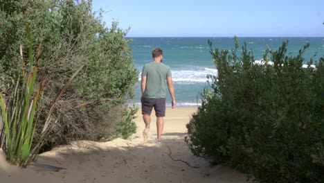 Male-walking-barefoot-down-sandy-track-towards-windy-beach-ocean-waves