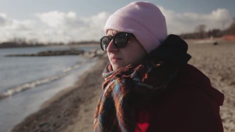 Young-Tourist-Wearing-Red-Jacket,-Sun-glasses-and-Pink-beanie-hat-Standing-On-The-Shore-and-Enjoying-The-Bright-Summer-Day---Close-Up-Shot