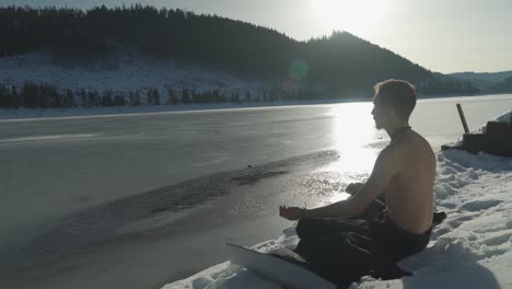 Man-meditating-on-mat-sitting-in-snow-beside-frozen-mountain-lake-with-sun-on-horizon-and-sunlight-shining-on-ice-covered-surface