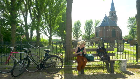 A-female-cyclist-resting-on-a-bench-drinking-some-water-in-front-of-a-church-in-Friesland,-zurich