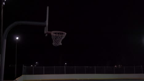 Teenage-girl-kicks-basketball-up-with-foot-and-shoots-it-in-the-net-at-dark-outdoor-court-with-lights-and-rain