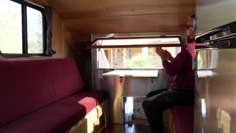 A-man-opening-the-window-in-slow-motion-inside-of-a-deluxe-custom-teardrop-travel-trailer-in-a-sunny-forest-campsite