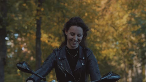 Pretty-smiling-European-young-woman-driving-a-motorbike-wearing-leather-jacket-in-forest-with-vibrant,-colorful-golden-autumn-leaves-on-sunny-day-5