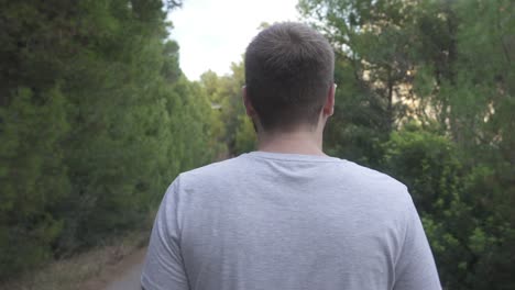 The-back-of-a-man-dressed-in-basic-gray-T-shirt,-who-walks-along-a-rural-road-surrounded-by-nature-and-trees-on-the-sides-of-the-image
