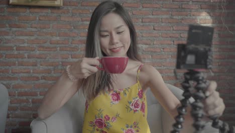 Beautiful-asian-travel-influencer-in-vibrant-yellow-spaghetti-top-holds-tripod-to-record-herself,-smiles-at-camera,-talks-to-camera-and-sips-coffee-on-a-light-brown-couch-in-a-warm-tone-cafe