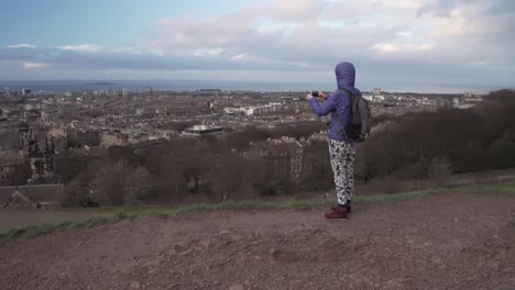 Tracking-follow-shot-of-a-girl-walking-to-the-edge-of-a-cliff-taking-a-picture-with-smartphone-overlooking-the-whole-city-of-Edinburgh-at-sunset-from-Calton-hill,-Scotland