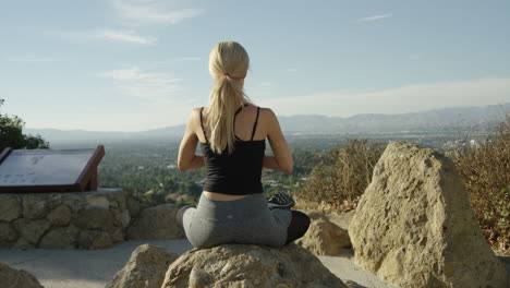 SLOW-MOTION-video-of-young-blonde-woman-seen-from-behind-sitting-on-a-rock-outside-in-yoga-pose-with-blue-sky-and-beautiful-background