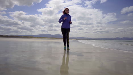 Low-dolly-shot-of-a-girl-running,-jogging-on-the-shore-of-a-sandy-beach-with-Atlantic-ocean-waves-on-a-wonderful-sunny-day-in-Ireland-in-4K