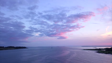 Aerial-shot-flying-out-over-a-calm-Atlantic-Ocean-harbor-with-a-single-sailboat-during-a-colorful-pink-and-purple-sunset-off-the-Maine-coast