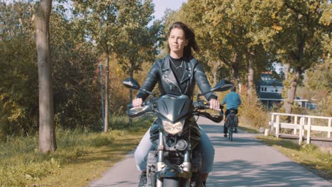 Front-view-of-beautiful-smiling-European-Female-motorcyclist-wearing-leather-driving-On-road-passing-cyclists-with-autumn-leaf-color-Trees-in-forest-on-sunny-day