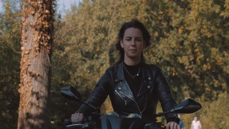 Front-perspective-of-pretty-smiling-European-young-lady-driving-a-motorbike-wearing-leather-jacket-in-forest-with-vibrant,-colorful-golden-autumn-leaves-on-sunny-day