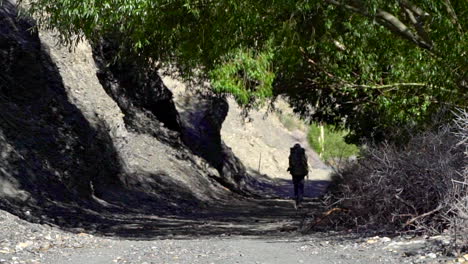 Man-with-backpack-walking-away-in-the-shadows-on-a-sunny-day-on-a-dirt-road-in-the-mountains