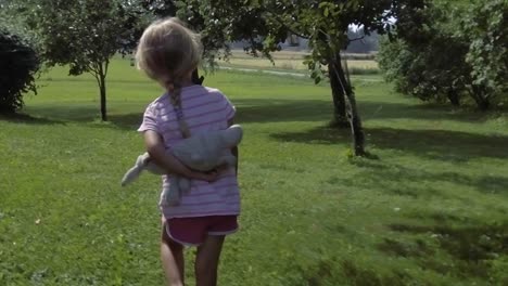 Three-year-old-summer-girl-walking-on-grass-field-among-apple-trees,-holding-stuffed-animal,-pink-clothes,-slow-motion
