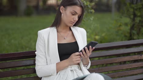 Young-business-woman-checks-her-smartphone,-sitting-on-a-bench-in-the-park