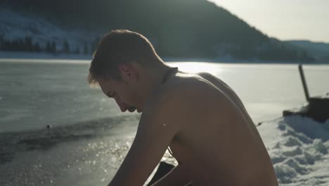 Man-with-no-shirt-sitting-in-snow-beside-frozen-lake