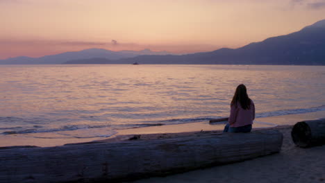 Eine-Junge-Frau-Sitzt-Und-Beobachtet-Nachdenklich-Den-Wunderschönen-Sonnenuntergang-An-Einem-Ruhigen-Strand-Mit-Blick-Auf-Die-Berge-Im-Wunderschönen-British-Columbia,-Kanada