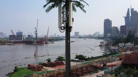 Ascending-aerial-shot-of-ultra-modern-mobile-phone-tower-disguised-as-a-palm-tree-against-a-background-of-a-riverfront-construction-site-with-city-buildings,-cranes-and-boat-traffic-on-the-river
