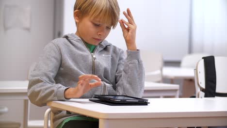 Schoolboy-in-classroom-at-desk-studying-with-tablet,-Pan-medium-shot