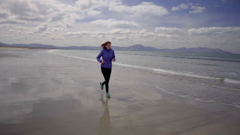 Tracking-dolly-shot-of-a-girl-running,-jogging-on-the-shore-of-a-sandy-beach-with-Atlantic-ocean-waves-on-a-wonderful-sunny-day-in-Ireland-in-4K