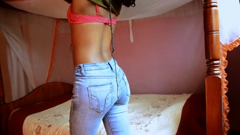 African-woman-taking-off-cropped-belly-shirt-revealing-pink-bra-wearing-tight-jeans-in-front-of-a-bed-in-a-hotel-room---SIDE-VIEW
