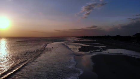 Unglaubliche-Luftaufnahme-Eines-Farbenfrohen-Sonnenuntergangs-An-Einem-Sandstrand,-Menschen,-Die-Am-Strand-Spielen-–-Vom-Strand-Aus-Seitlich-In-Richtung-Meer-Fliegen