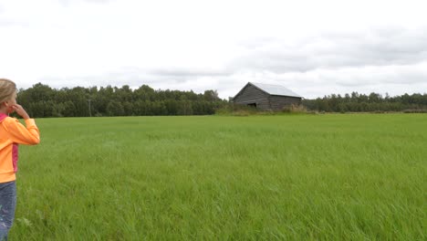 Freedom-of-Summer-break,-Alone-blond-young-girl-walking-on-grass-field,-approaching-old-barn,-Slow-motion