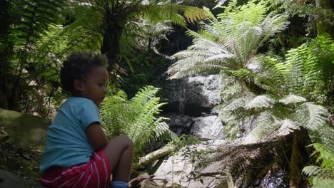A-young-child-sits-on-a-ledge-in-a-rain-forest-looking-at-a-small-waterfall