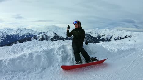 Snowboarder-Adolescente-Tomando-Selfie-Sentado-En-Una-Nieve-Rodeada-De-Montañas
