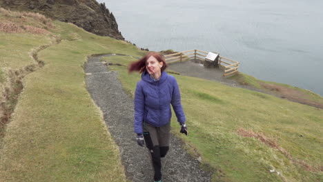 Follow-shot-of-happy-girl-walking-on-the-edge-of-cliffs-in-Scotland,-isle-of-Skye-with-Atlantic-Ocean-in-the-background-on-a-cloudy-day