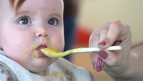 A-close-up-shot-of-a-mothers-hand-nudging-a-spoon-full-of-baby-food-into-a-toddlers-mouth,-then-the-toddler-becomes-unhappy-and-spits-it-out,-close-up-shot-in-slow-motion