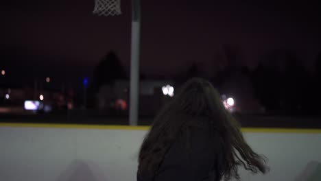 Girl-with-brown-hair-runs-up-and-shoots-layup-into-net-at-outdoor-basketball-court-with-lights-at-night