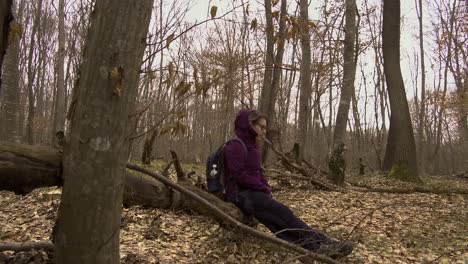 Woman-in-purple-jacket-with-backpack-and-water-bottle-sitting-on-a-fallen-tree-in-the-forest
