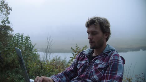 Medium-shot,-young-man-using-a-laptop-while-looking-around,-foggy-lake-view-in-the-background