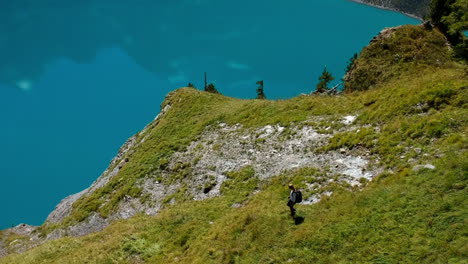 Arial-shot-of-a-blonde-woman-with-backpack-hiking-down-a-lush-mountain-at-a-blue-mountain-lake-in-the-swiss-alps
