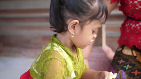 Entzückendes-Kleines-Indonesisches-Mädchen-Stellt-Eine-Blumendekoration-Aus-Streifen-Von-Palmblättern-Her-Und-Heftet-Das-Endprodukt