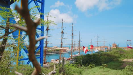 Langsamer-Schwenk,-Der-Eine-Regatta-Von-Großseglern-Zeigt,-Die-Entlang-Des-Flusses-Mit-Einer-Stadt-Und-Einer-Blauen-Strukturellen-Unterstützung-Für-Eine-Brücke-In-Curaçao-Festgemacht-Sind