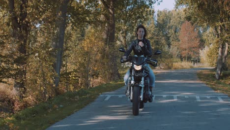 Pretty-smiling-European-young-woman-driving-a-motorbike-wearing-leather-jacket-in-forest-with-vibrant,-colorful-golden-autumn-leaves-on-sunny-day-6