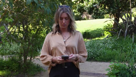 Young-woman-walking-through-garden-on-her-mobile-phone_slow-motion-1