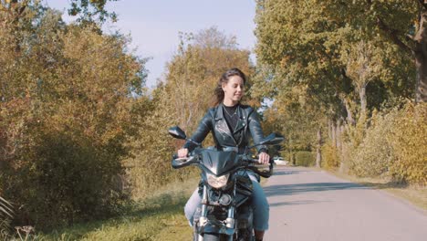 Lächelnde-Junge-Dame-Auf-Dem-Motorrad,-Die-An-Einem-Sonnigen-Tag-Durch-Wald-Und-Ländliche-Felder-Mit-Goldenen-Herbstlaubbäumen-Fährt